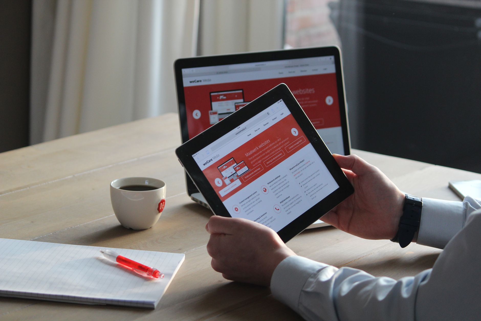 Pessoa segurando um tablet com um site aberto, com um notebook e uma xícara de café sobre a mesa ao fundo