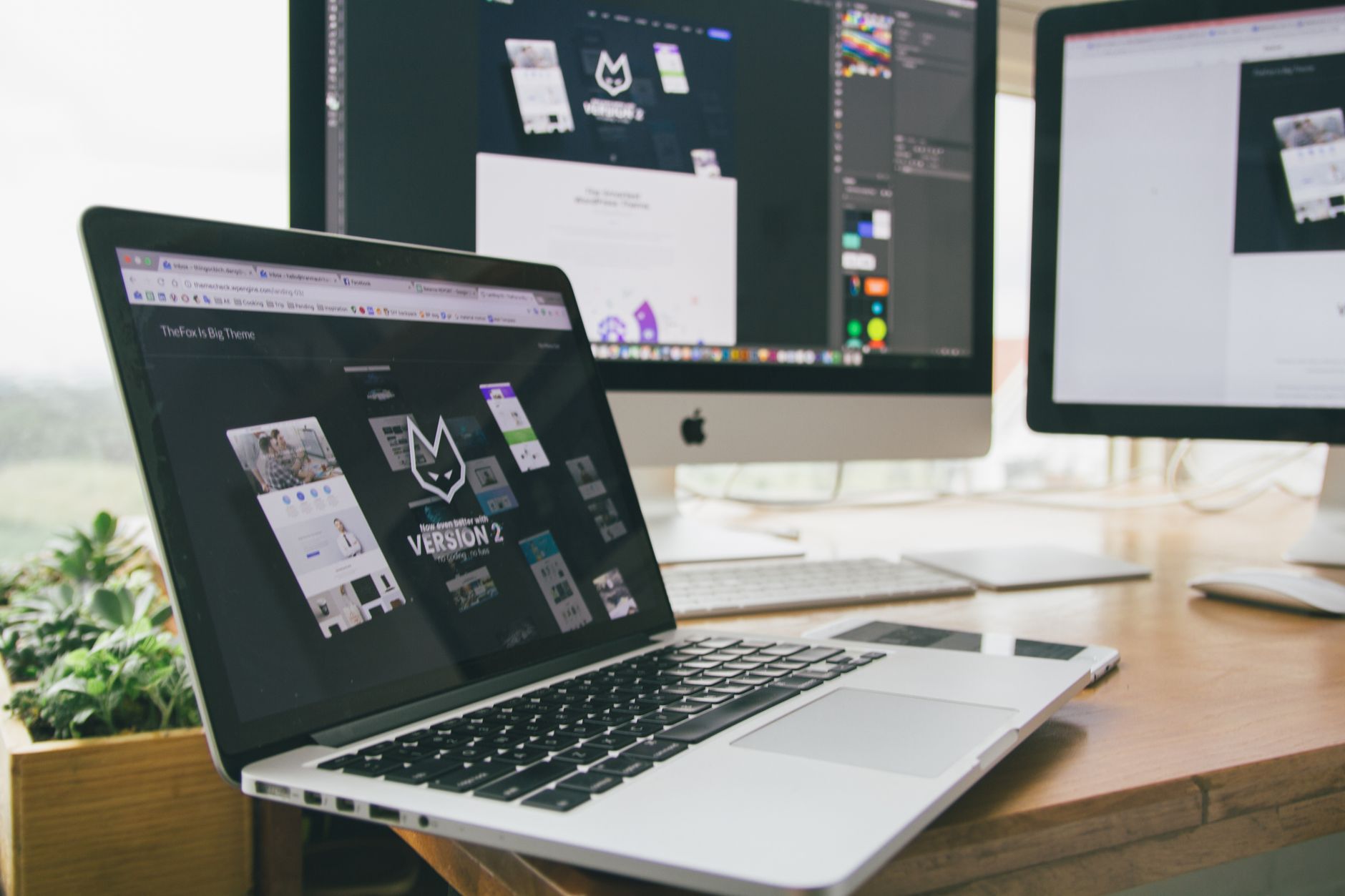 Notebook aberto sobre mesa de escritório, com monitores de computador ao fundo exibindo telas de design de site
