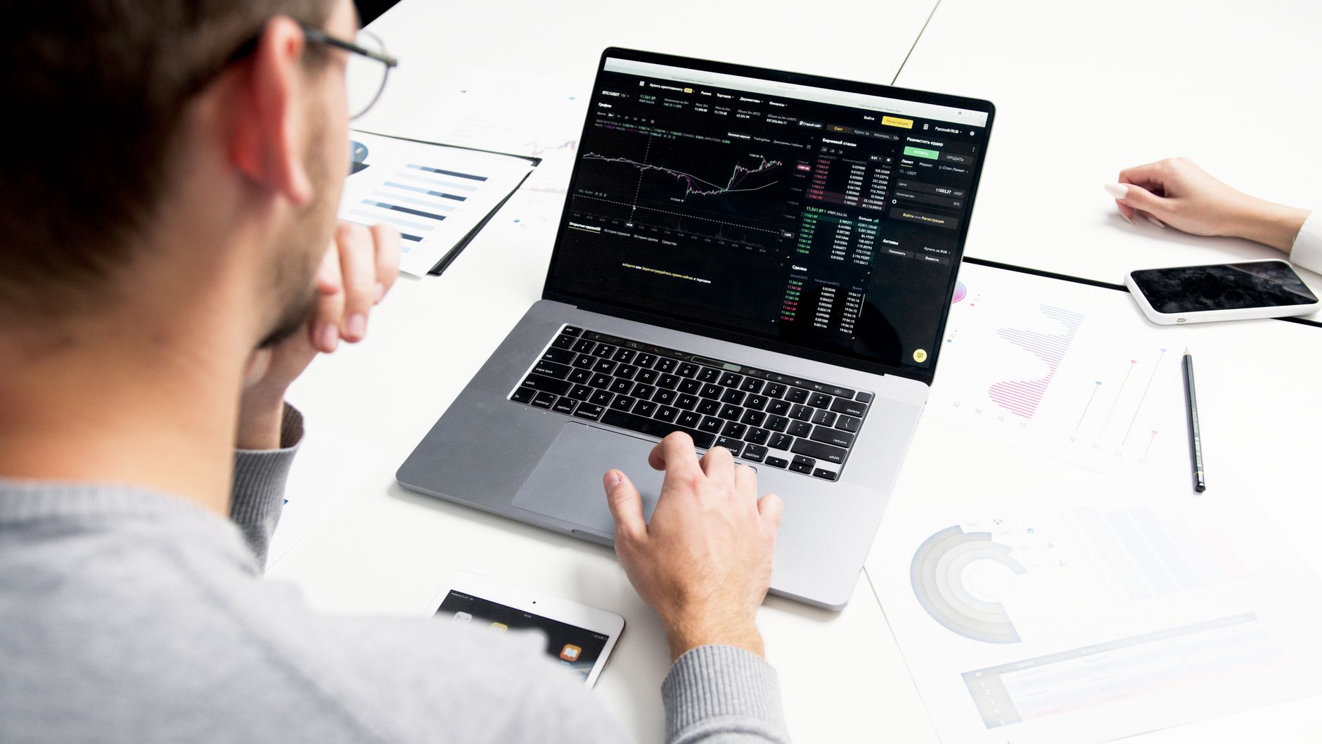 Pessoa analisando gráfico financeiro em laptop com papéis contendo gráficos ao redor e celular ao lado