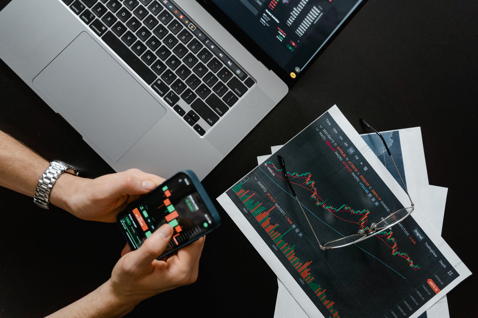 Pessoa segurando smartphone com gráficos ao lado de notebook e relatórios impressos com gráficos financeiros