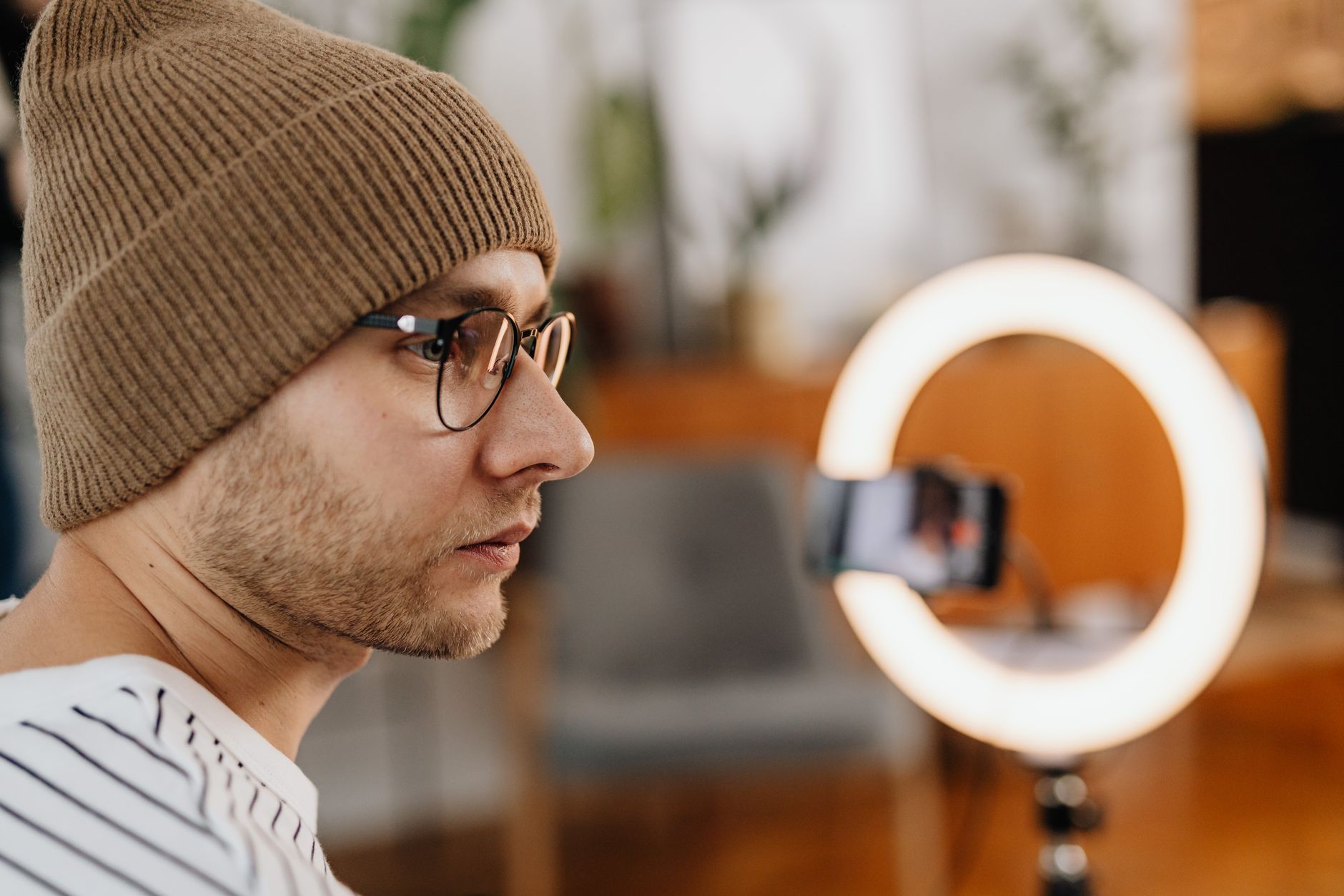 Close-up de perfil de homem de touca e óculos conversando em frente a câmera de celular com luz de anel ao fundo