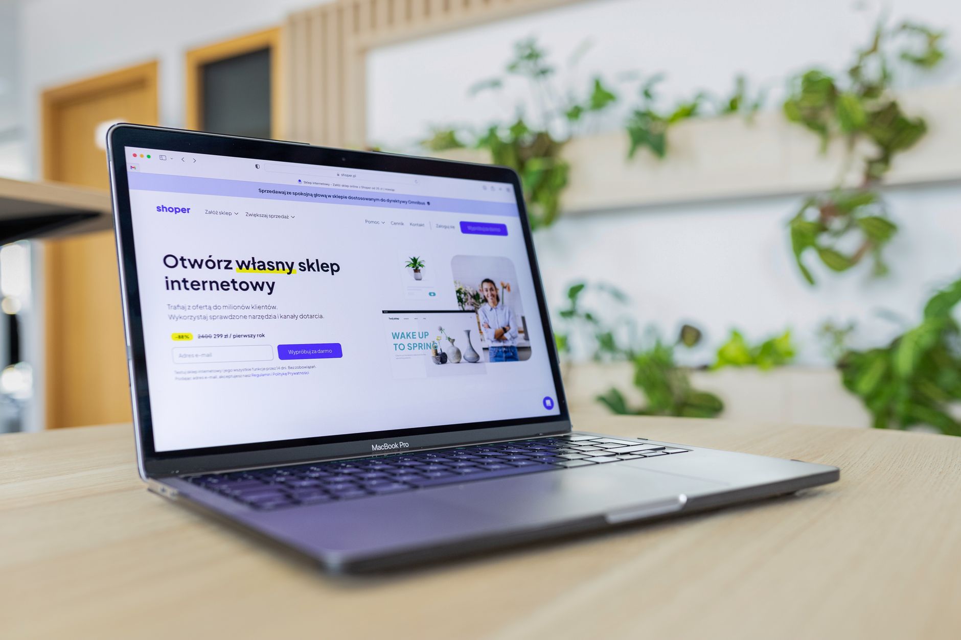 Laptop sobre mesa de madeira exibindo a página inicial de uma plataforma de loja virtual, com plantas ao fundo
