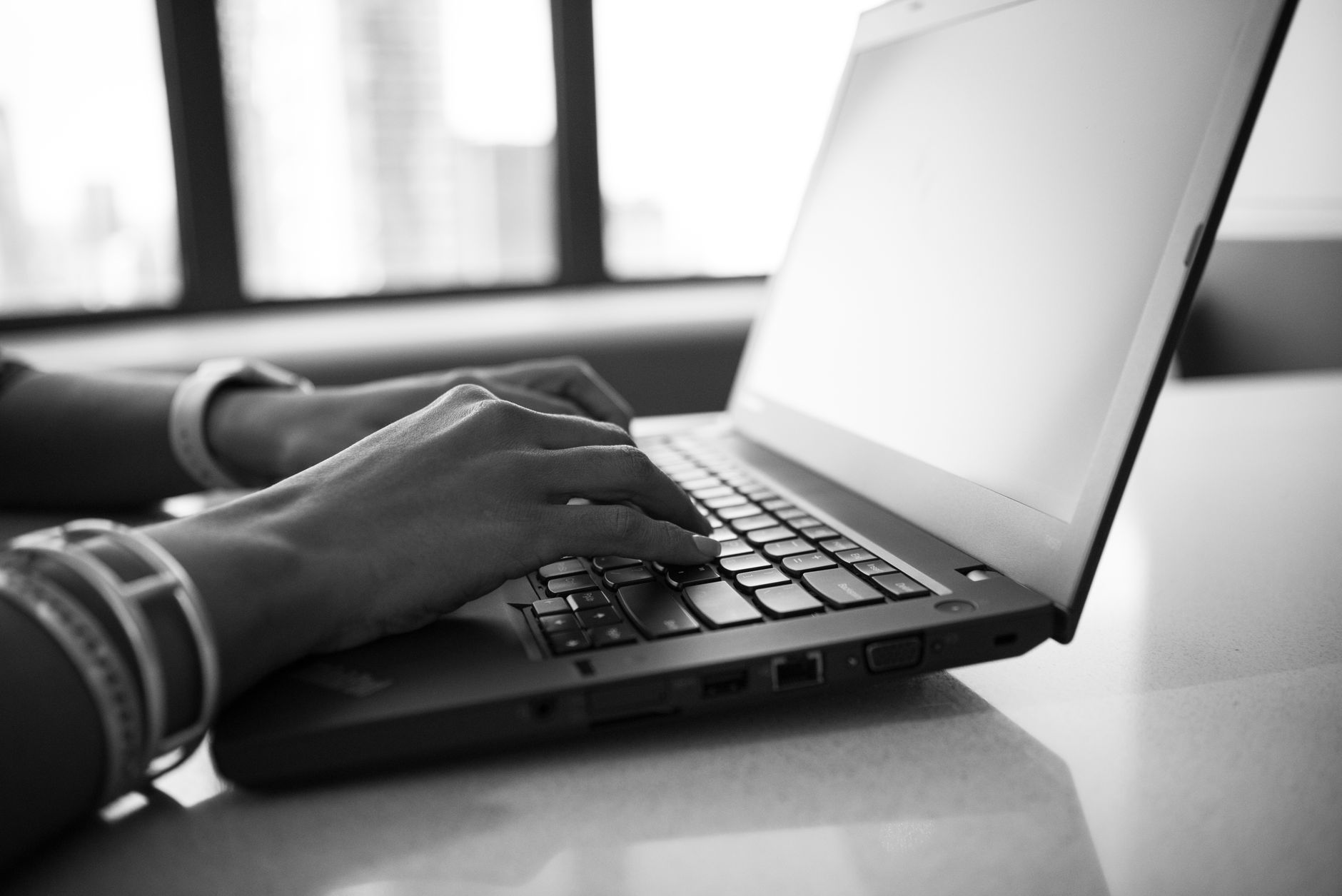 Mãos digitando em um notebook em preto e branco, próximo a uma janela